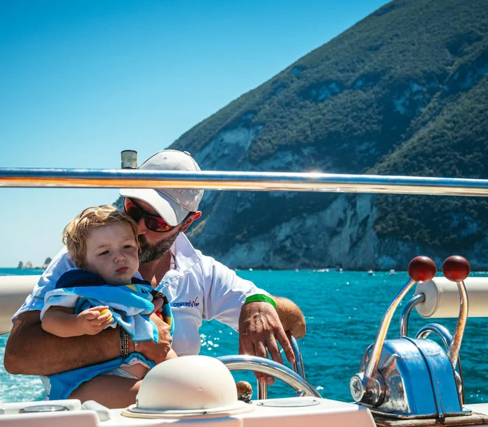 Escursioni in barca al Conero e molto altro Uomo con bambino al timone di una barca sul mare con colline sullo sfondo