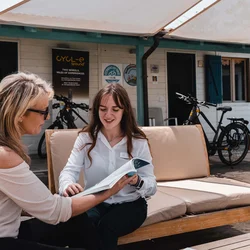 Le migliori offerte per le vostre vacanze al mare Due donne sedute su un divano all'aperto consultano un catalogo davanti a un negozio di biciclette