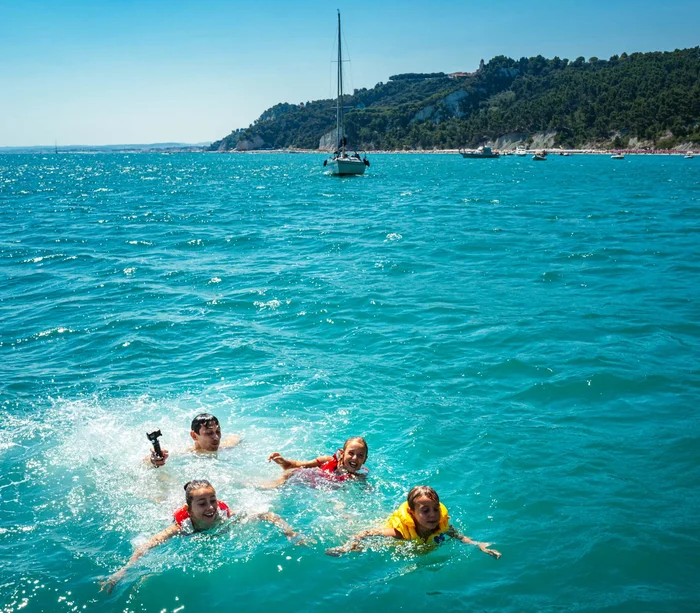 Escursioni in barca al Conero e molto altro Quattro bambini che nuotano nel mare con una barca a vela sullo sfondo