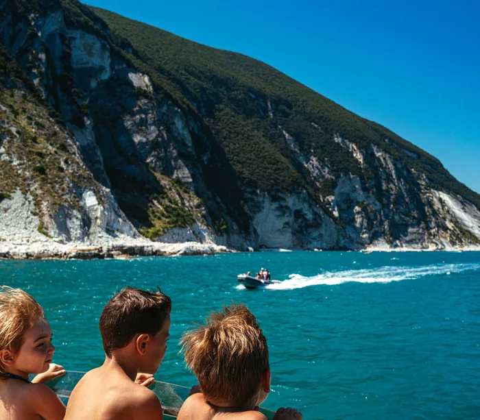 Una vacanza nelle Marche: terra di emozioni Tre bambini su una barca guardano il mare e una barca veloce vicino alla costa rocciosa