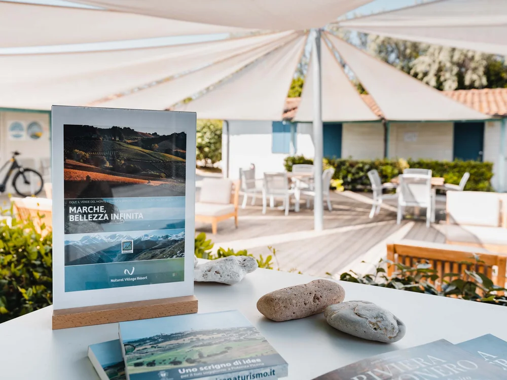 Resort al mare: pronti per un’escursione sul Conero? Volantini promozionali su un tavolo all'aperto con sedie e ombrelloni bianchi sullo sfondo