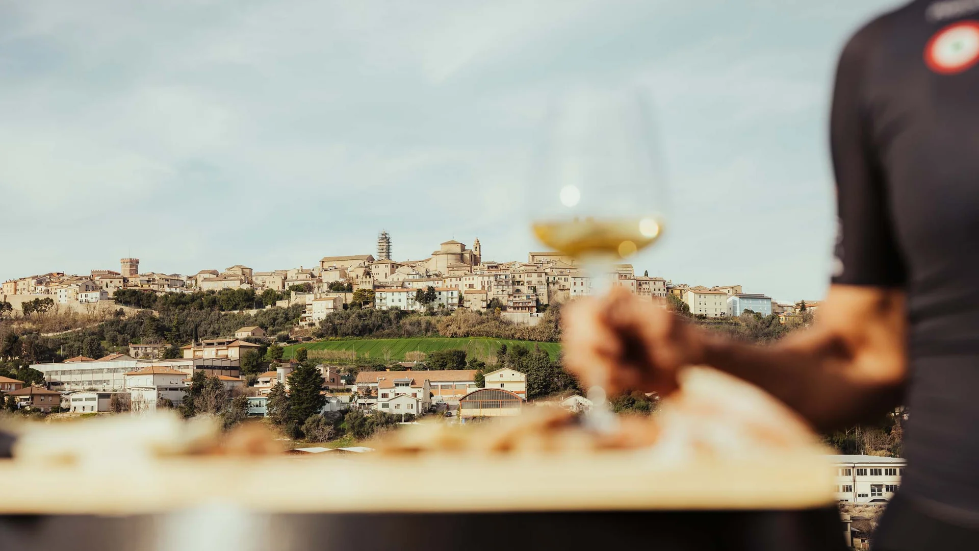 Borghi delle Marche: un trionfo di arte e cultura Vista di un borgo collinare con una mano che tiene un calice di vino bianco