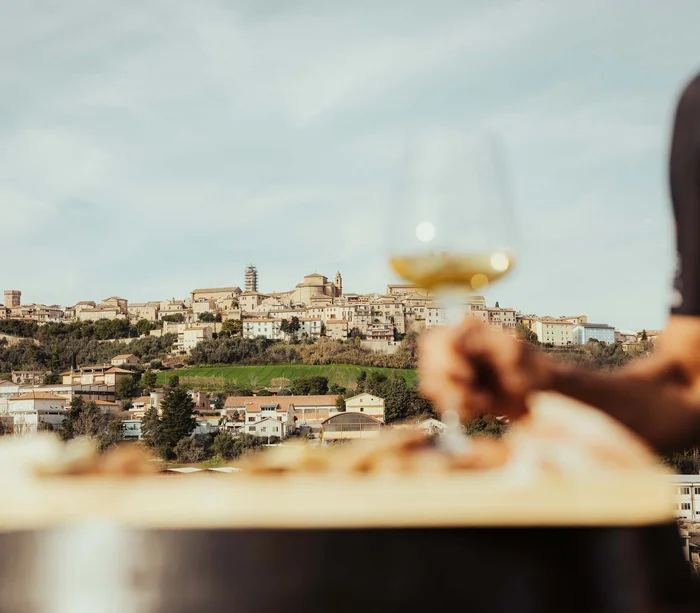 Una vacanza nelle Marche: terra di emozioni Vista di un borgo collinare con una mano che tiene un calice di vino bianco
