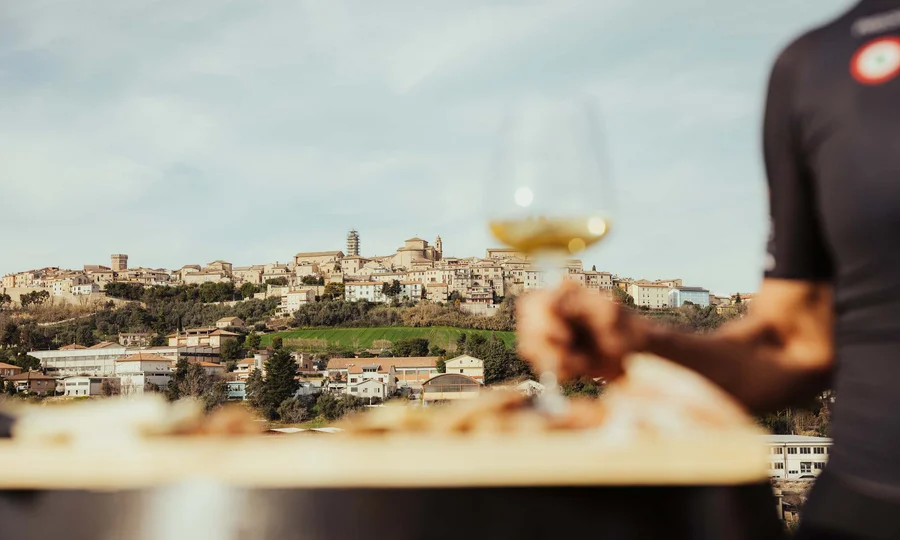 Resort al mare: pronti per un’escursione sul Conero? Vista di un borgo collinare con una mano che tiene un calice di vino bianco
