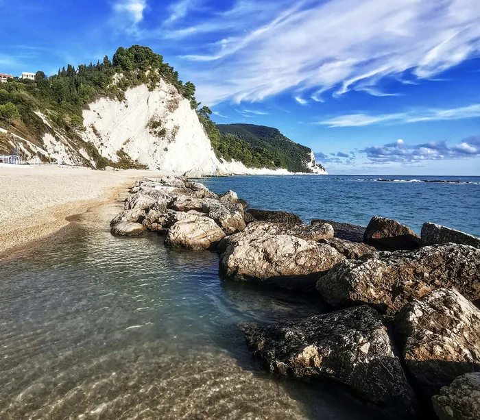Una vacanza nelle Marche: terra di emozioni Spiaggia rocciosa con acqua trasparente e cielo azzurro con nuvole