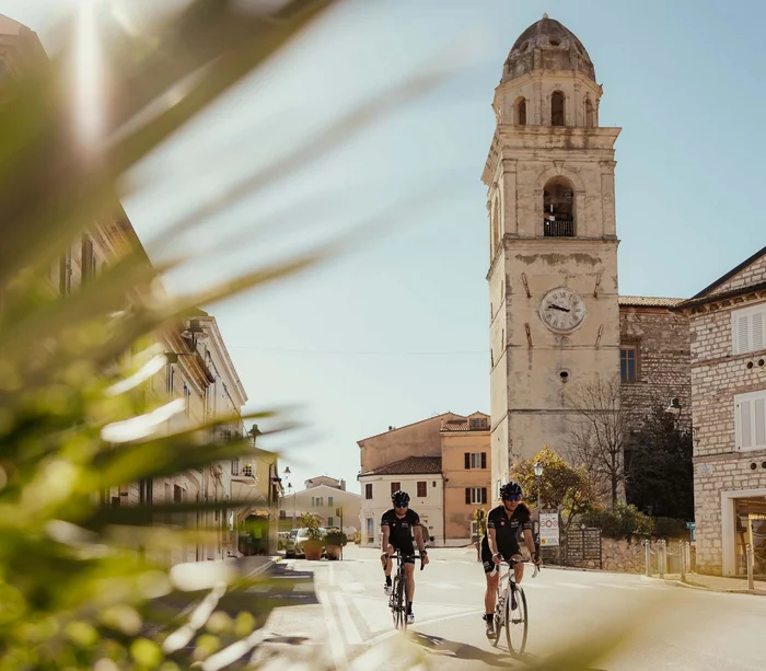 Una vacanza nelle Marche: terra di emozioni Due ciclisti su bici da corsa in una piazza con torre dell'orologio