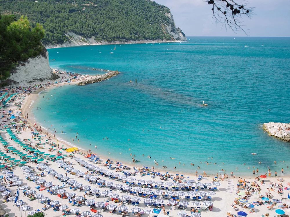 Spiaggia Urbani: la mezzaluna di Sirolo Spiaggia affollata con ombrelloni e mare turchese con scogliere verdi