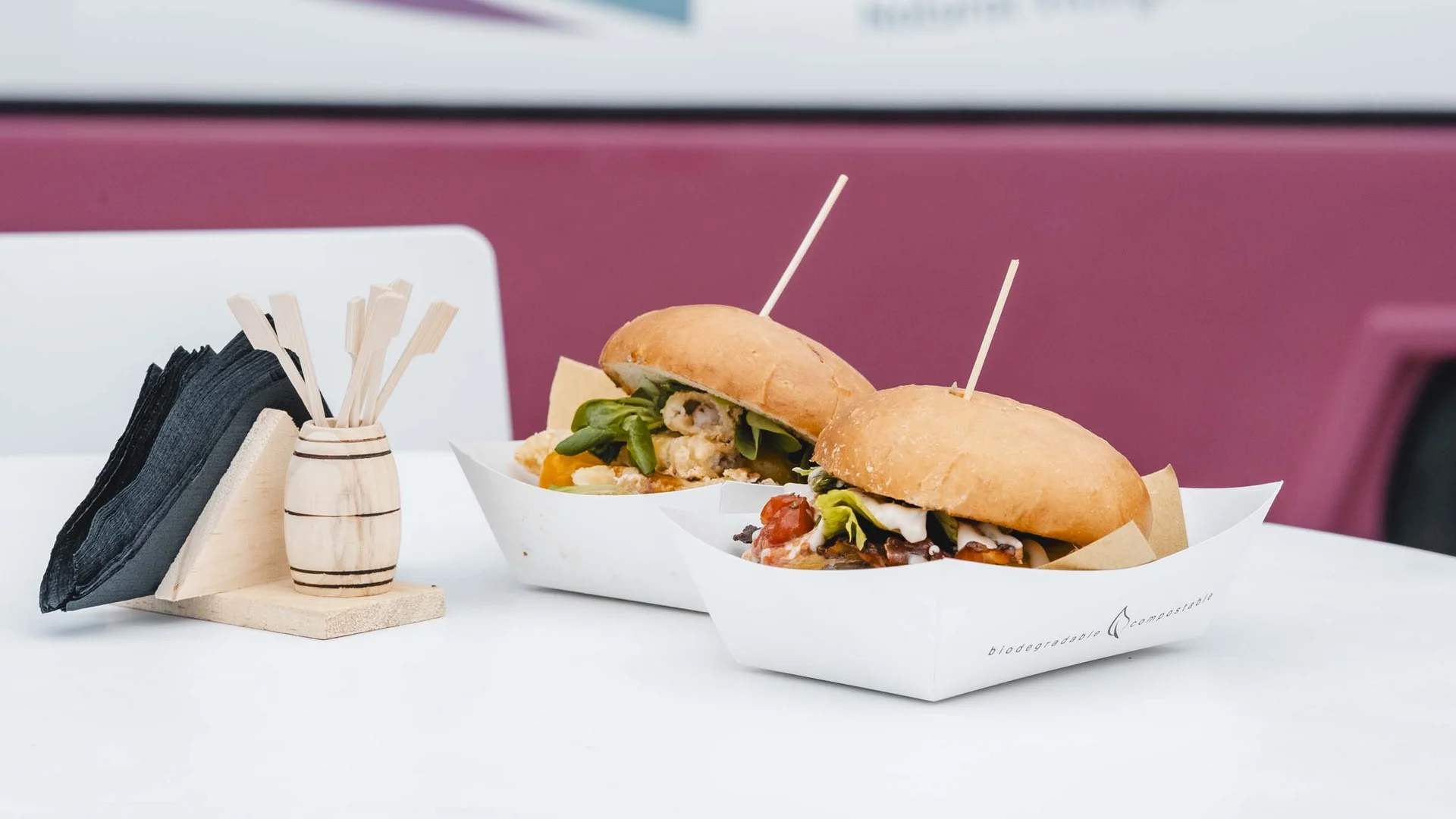 Food truck & Pizzeria a Potenza Picena Due hamburger gourmet in vassoi di carta su tavolo bianco con tovaglioli e food truck sullo sfondo