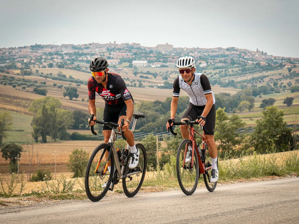 Signature Experience Due ciclisti in abbigliamento da corsa pedalano su strada di campagna con paese sullo sfondo