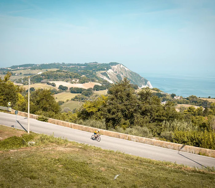 Una vacanza nelle Marche: terra di emozioni Ciclista su strada di montagna con vista sul mare e colline verdi