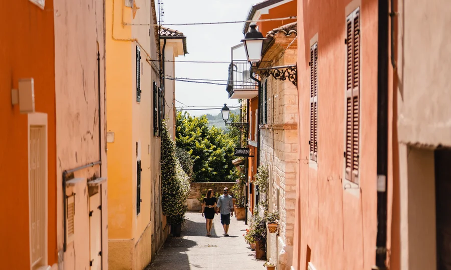 Resort al mare: pronti per un’escursione sul Conero? Coppia che cammina in una stretta strada di un villaggio con case colorate