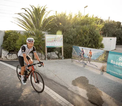 Un family hotel al Conero? Meglio un resort! Ciclista su strada vicino a cartello pubblicitario Roadbike Holidays