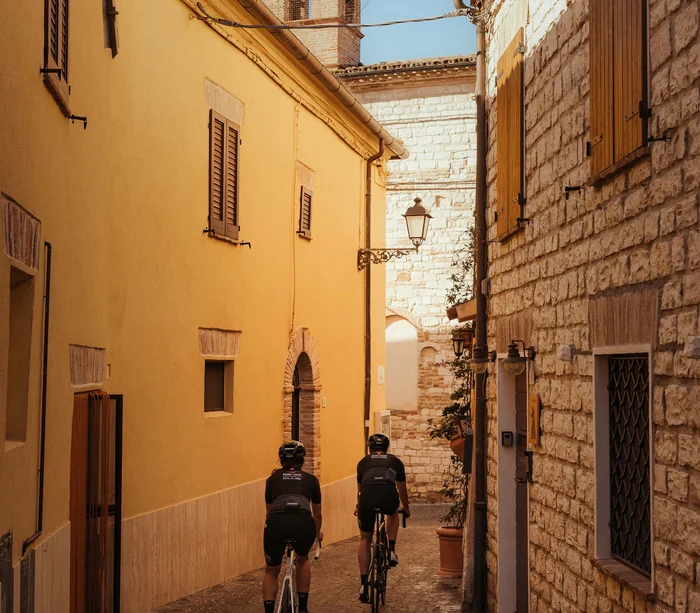 Una vacanza nelle Marche: terra di emozioni Due ciclisti che pedalano in una stretta via tra vecchi edifici in pietra