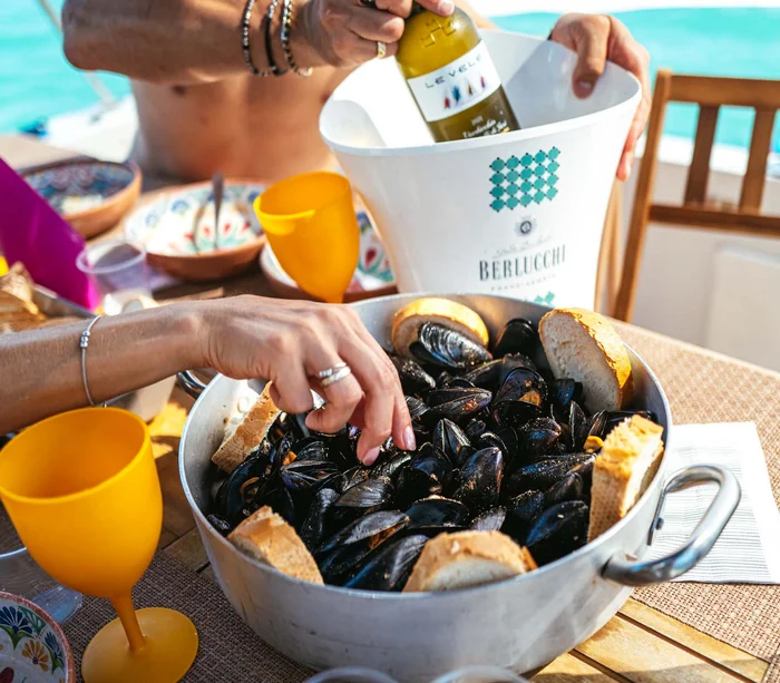Escursioni in barca al Conero e molto altro Mano che prende cozze da una pentola con pane e bicchieri gialli sul tavolo