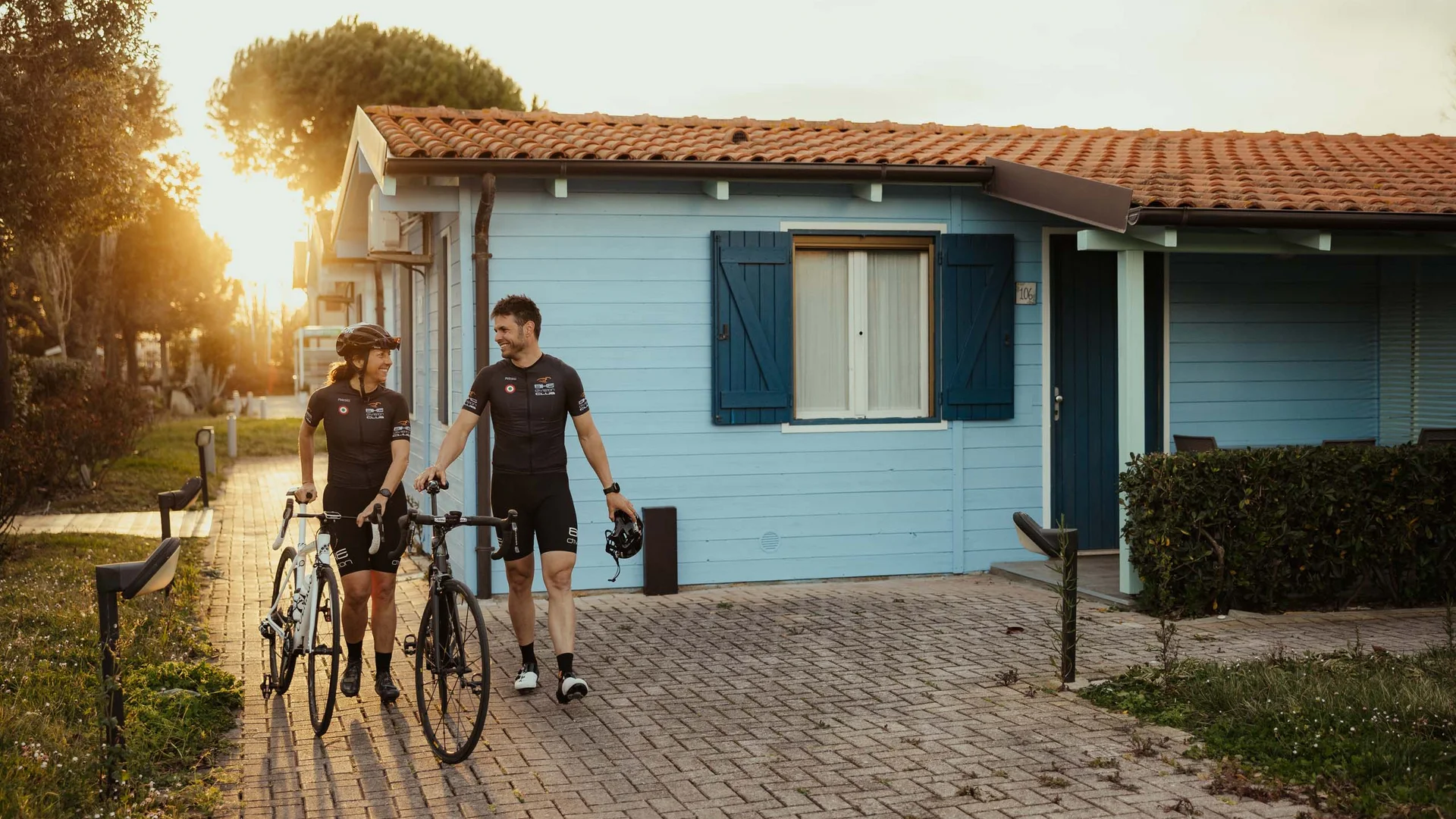Un villaggio nelle Marche sul mare: il Natural Village Resort Due ciclisti vestiti in abbigliamento tecnico camminano accanto a una casa azzurra al tramonto