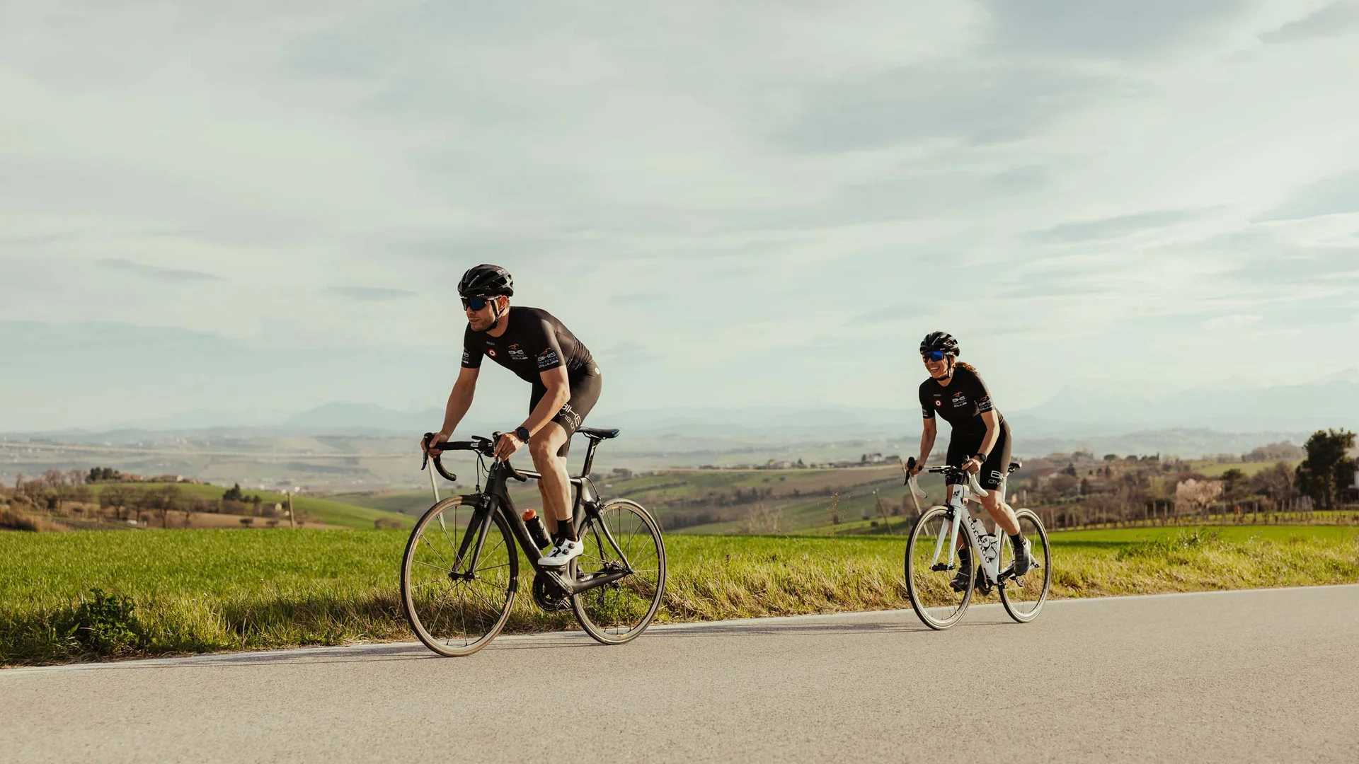 What could be better than a bike hotel in the Marche region? Two cyclists in gear riding on paved road through countryside.