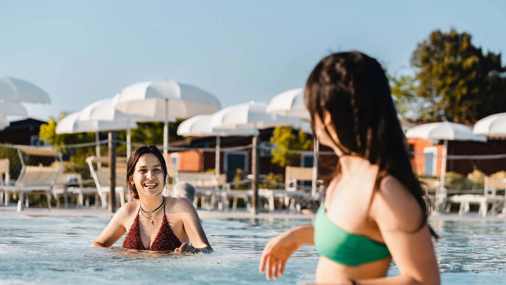 Un villaggio nelle Marche sul mare: il Natural Village Resort Due donne sorridenti in costume da bagno in piscina con ombrelloni bianchi sullo sfondo